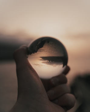 A hand holding a crystal ball that reflects an upside-down image of a landscape at sunset, with a cloudy sky and a distant landmass across a body of water. The background appears blurred, accentuating the clarity within the crystal ball.
