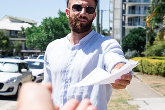 A man wearing sunglasses and a light blue shirt is extending a document towards the viewer while standing outdoors. Cars are parked on the side, and buildings and greenery are visible in the background.