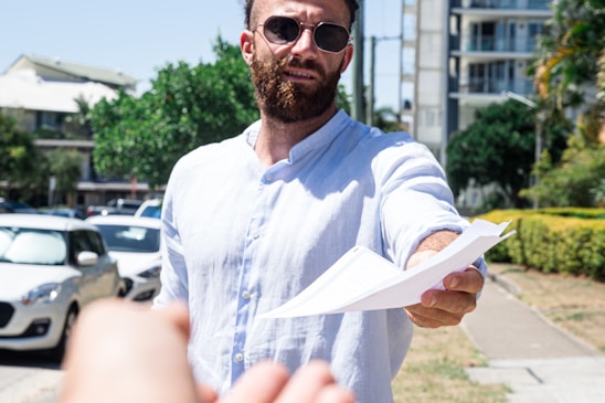 A man wearing sunglasses and a light blue shirt is extending a document towards the viewer while standing outdoors. Cars are parked on the side, and buildings and greenery are visible in the background.