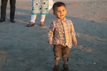 A young child stands on a sandy surface, dressed in a plaid shirt and rolled-up jeans. The child is smiling and looking towards the camera. In the background, the legs of two adults are visible, partially showing their attire. The scene is warmly lit, suggesting it is either morning or late afternoon.