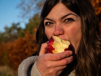 A woman is eating a red apple, biting into it with a look of satisfaction or determination. She is wearing a gray knitted sweater, and her long dark hair is partially visible. The background features blurred autumn foliage with warm tones and a clear blue sky.