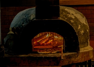 A rustic stone oven is illuminated by the warm glow of fire inside. Several pizzas are cooking on the stone surface, with flames visibly heating the interior. The oven's exterior has a weathered look, surrounded by a brick wall.