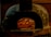 A rustic stone oven is illuminated by the warm glow of fire inside. Several pizzas are cooking on the stone surface, with flames visibly heating the interior. The oven's exterior has a weathered look, surrounded by a brick wall.