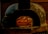 A rustic stone oven is illuminated by the warm glow of fire inside. Several pizzas are cooking on the stone surface, with flames visibly heating the interior. The oven's exterior has a weathered look, surrounded by a brick wall.
