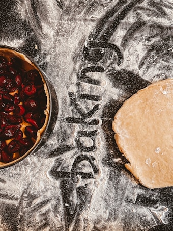 Flour scattered across a surface with the word 'baking' written in it. A pie crust filled with cherries is on the left and a rolled out dough is on the right.