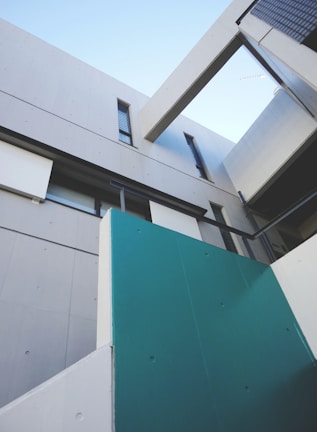 A modern architectural structure with clean, sharp lines and a minimalist design. The exterior features concrete walls and large geometric panels in grey and teal. Angled beams and windows add to the contemporary aesthetic, with a clear blue sky visible above.