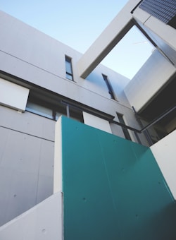 A modern architectural structure with clean, sharp lines and a minimalist design. The exterior features concrete walls and large geometric panels in grey and teal. Angled beams and windows add to the contemporary aesthetic, with a clear blue sky visible above.