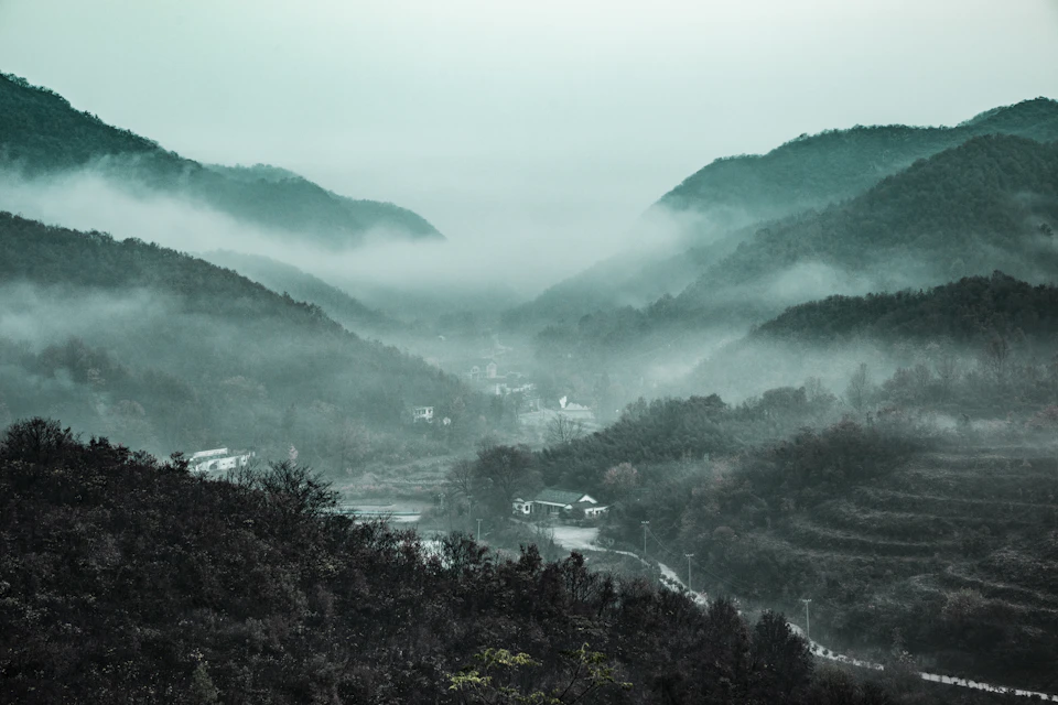 The image depicts a serene mountainous landscape shrouded in fog, with a small village nestled at the foot of the mountains. The village is surrounded by lush green trees and shrubs, creating a sense of tranquility. The perspective of the photo is from a high vantage point, providing a panoramic view of the village and its natural surroundings.