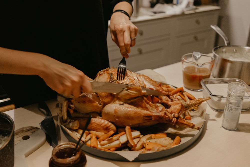 person holding stainless steel fork and bread knife slicing grilled meat on white ceramic plate