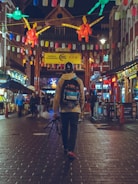 A person with a backpack is walking down a vibrant, lantern-lit street in a bustling city. The street is adorned with colorful decorations and signs, surrounded by various shops and eateries. Other people are seen in the background, adding to the lively atmosphere.