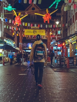 A person with a backpack is walking down a vibrant, lantern-lit street in a bustling city. The street is adorned with colorful decorations and signs, surrounded by various shops and eateries. Other people are seen in the background, adding to the lively atmosphere.