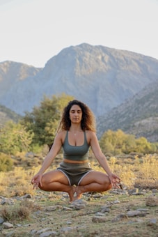 A person is meditating outdoors in a seated position, surrounded by a natural landscape with mountains and trees.