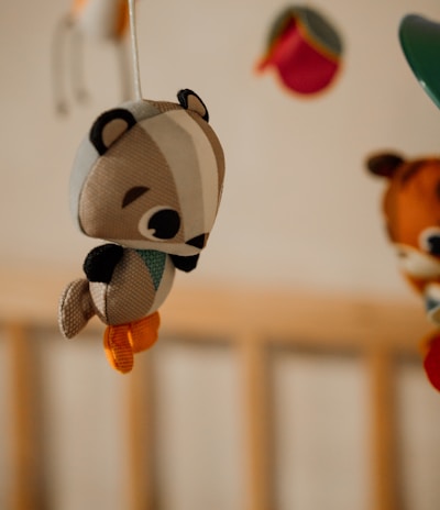 A soft plush toy resembling a small woodland animal, possibly a chipmunk or squirrel, hangs from a mobile. The toy is crafted with fabric in various colors including brown, white, orange, and green. In the background, other plush toys are partially visible, hanging from the same mobile structure, and a wooden crib frame can be seen.