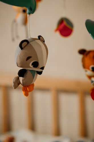 A soft plush toy resembling a small woodland animal, possibly a chipmunk or squirrel, hangs from a mobile. The toy is crafted with fabric in various colors including brown, white, orange, and green. In the background, other plush toys are partially visible, hanging from the same mobile structure, and a wooden crib frame can be seen.