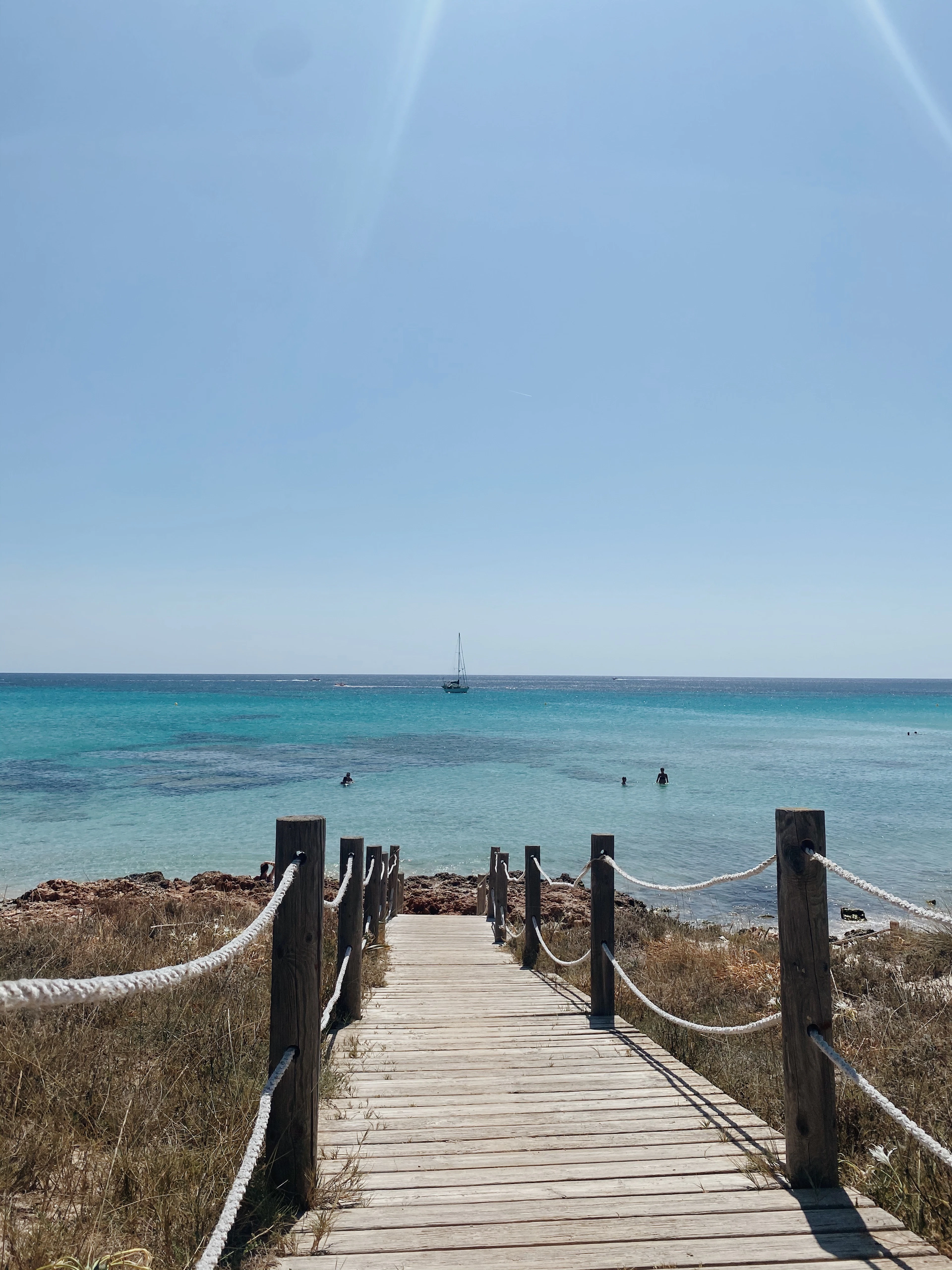 A rope-railed wooden boardwalk leads to a turquoise cove with a moored sailboat and swimmers off the Ibiza coast.