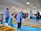 A group of people wearing protective clothing and caps stand in an indoor facility with blue and yellow fish tanks. Some individuals are taking notes while another person handles a net inside a tank. Overhead lights illuminate the room, and a banner with text is visible on the wall.