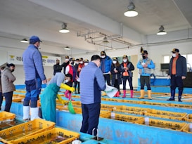 A group of people wearing protective clothing and caps stand in an indoor facility with blue and yellow fish tanks. Some individuals are taking notes while another person handles a net inside a tank. Overhead lights illuminate the room, and a banner with text is visible on the wall.