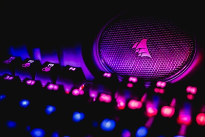 A close-up view of an illuminated gaming keyboard with vibrant LED lights and a speaker with a logo. The keys are lit in shades of pink and blue, creating a neon glow against a dark background.
