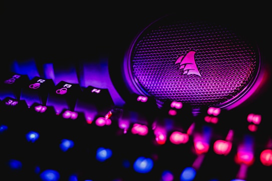 A close-up view of an illuminated gaming keyboard with vibrant LED lights and a speaker with a logo. The keys are lit in shades of pink and blue, creating a neon glow against a dark background.