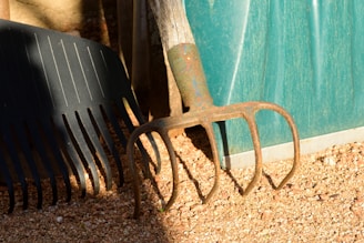 A weathered garden fork and a rake rest against a blue plastic container on gravel. The tools show signs of rust and wear, suggesting heavy use. They are partially illuminated by natural sunlight, casting shadows on the ground.