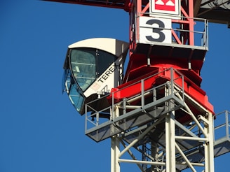 A construction crane's cabin sits atop a red and white steel structure with the number '3' prominently displayed. The crane cabin has large glass windows, and the sky in the background is clear and blue.