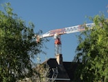 A large construction crane towers above a building with a dark-tiled roof surrounded by lush green trees. The clear blue sky forms the backdrop, highlighting the crane's red and white structure.
