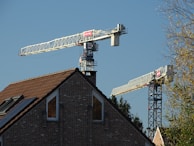 A brick house with a brown tiled roof is set against the backdrop of two large construction cranes. The cranes are positioned near the house, with the blue sky as the background. Trees are visible on the right side, adding a natural element to the predominantly industrial scene.