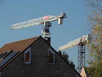 A brick house with a brown tiled roof is set against the backdrop of two large construction cranes. The cranes are positioned near the house, with the blue sky as the background. Trees are visible on the right side, adding a natural element to the predominantly industrial scene.