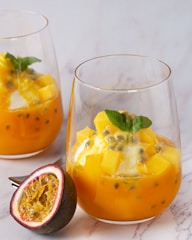 Two glasses of a vibrant orange dessert, possibly a fruit salad or parfait, containing chunks of yellow fruit, seeds, and garnished with a fresh green leaf. In the foreground, there is a halved passionfruit showing its juicy, seedy interior. The background is a simple, light-colored surface.