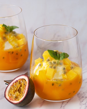 Two glasses of a vibrant orange dessert, possibly a fruit salad or parfait, containing chunks of yellow fruit, seeds, and garnished with a fresh green leaf. In the foreground, there is a halved passionfruit showing its juicy, seedy interior. The background is a simple, light-colored surface.