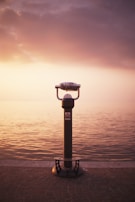 A public viewing telescope stands on a stone walkway overlooking a vast, calm body of water under a dramatic sunset. The sky is filled with warm hues of orange, pink, and purple, creating a serene and contemplative atmosphere.