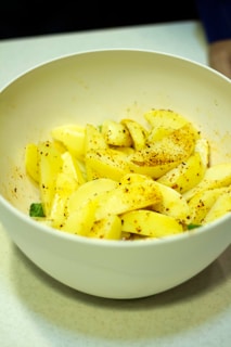 A white bowl contains sliced apples seasoned with spices. The apples are yellow and are sprinkled with a mix of red and brown spices, likely cinnamon or chili powder. The bowl is placed on a smooth, light-colored surface.
