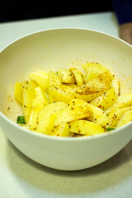A white bowl contains sliced apples seasoned with spices. The apples are yellow and are sprinkled with a mix of red and brown spices, likely cinnamon or chili powder. The bowl is placed on a smooth, light-colored surface.
