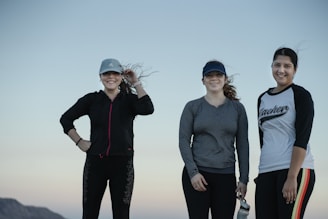 Three women stand outdoors against a clear sky, wearing casual athletic clothing and caps. They are smiling, suggesting a positive and friendly interaction. One holds a water bottle, indicating a possible outdoor activity or exercise.