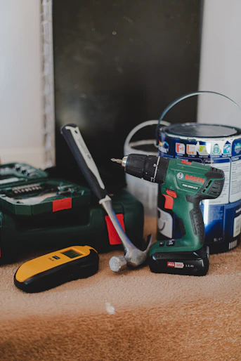 A cordless drill, a hammer, and a yellow stud finder on a carpeted floor
