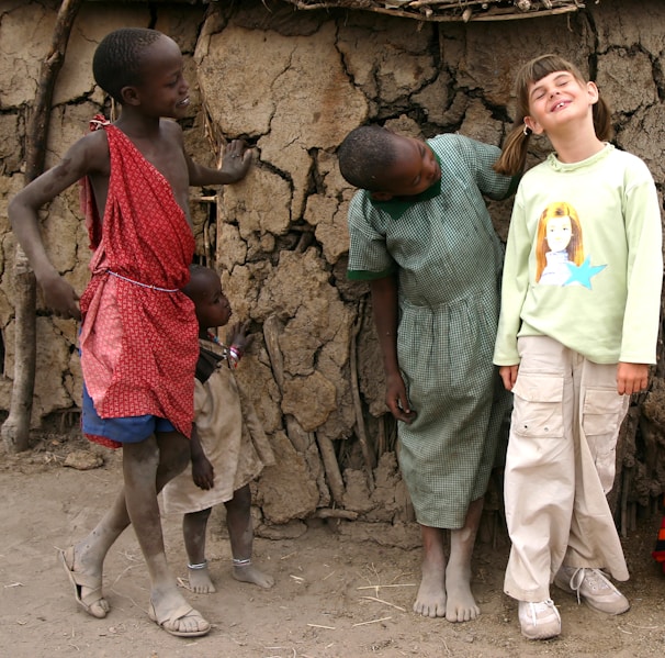 Four children are standing in front of a rustic mud wall. The two on the left are wearing traditional clothing, with one wearing a red patterned wrap. The child in the middle is in a green checkered dress, while another child in a green long-sleeve shirt with a cartoon illustration is smiling. The general atmosphere appears playful and friendly.