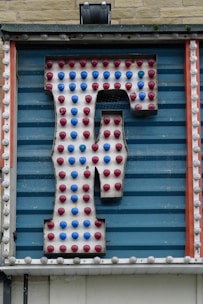 A large retro-style letter 'F' is attached to a striped blue background. The letter is adorned with alternating red and blue light bulbs, suggesting it might be part of a marquee or old sign. The structure is framed by vertical orange and white strips with additional circular detailing.