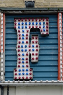 A large retro-style letter 'F' is attached to a striped blue background. The letter is adorned with alternating red and blue light bulbs, suggesting it might be part of a marquee or old sign. The structure is framed by vertical orange and white strips with additional circular detailing.