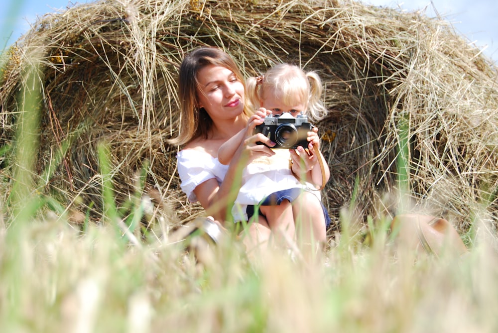 woman in white tank top holding black dslr camera sitting on brown grass field during daytime
