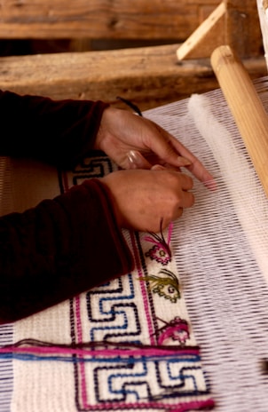 A person is weaving fabric on a loom. The hands are actively working with threads to create a pattern that includes intricate designs and colors. The background consists of wooden components of the weaving device, and the fabric being woven has detailed geometric and floral patterns.