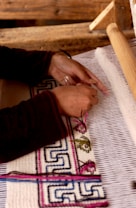 A person is weaving fabric on a loom. The hands are actively working with threads to create a pattern that includes intricate designs and colors. The background consists of wooden components of the weaving device, and the fabric being woven has detailed geometric and floral patterns.