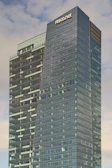 A modern skyscraper with a sleek glass façade towers against a cloudy sky. The building has a distinctive angular design with a prominent logo near the top of the structure. Windows cover the entire surface, providing a reflective quality that blends with the sky.