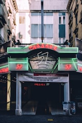 An urban passageway with neon signage above, displaying the words 'Passage 57' in red and green lights. The entrance, open and covered with a glass canopy, leads into a dark interior space. Above the entrance, there is a banner indicating available commercial space, with contact information. The surrounding buildings feature balconies and windows, indicating an architectural setting.
