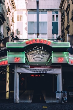 An urban passageway with neon signage above, displaying the words 'Passage 57' in red and green lights. The entrance, open and covered with a glass canopy, leads into a dark interior space. Above the entrance, there is a banner indicating available commercial space, with contact information. The surrounding buildings feature balconies and windows, indicating an architectural setting.
