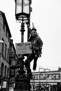 A person with a punk style, featuring a mohawk haircut and wearing a studded leather jacket, is perched on a lamppost in an urban setting. The individual holds a sign that reads 'Help a punk to get drunk £1 photo.' The background consists of brick buildings and a bridge with signage that suggests a city environment.