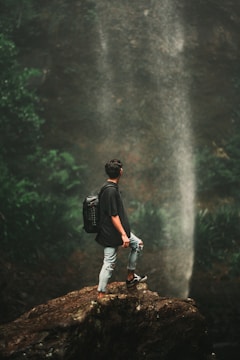 man in black t-shirt and blue denim jeans standing on rock near waterfalls during daytime