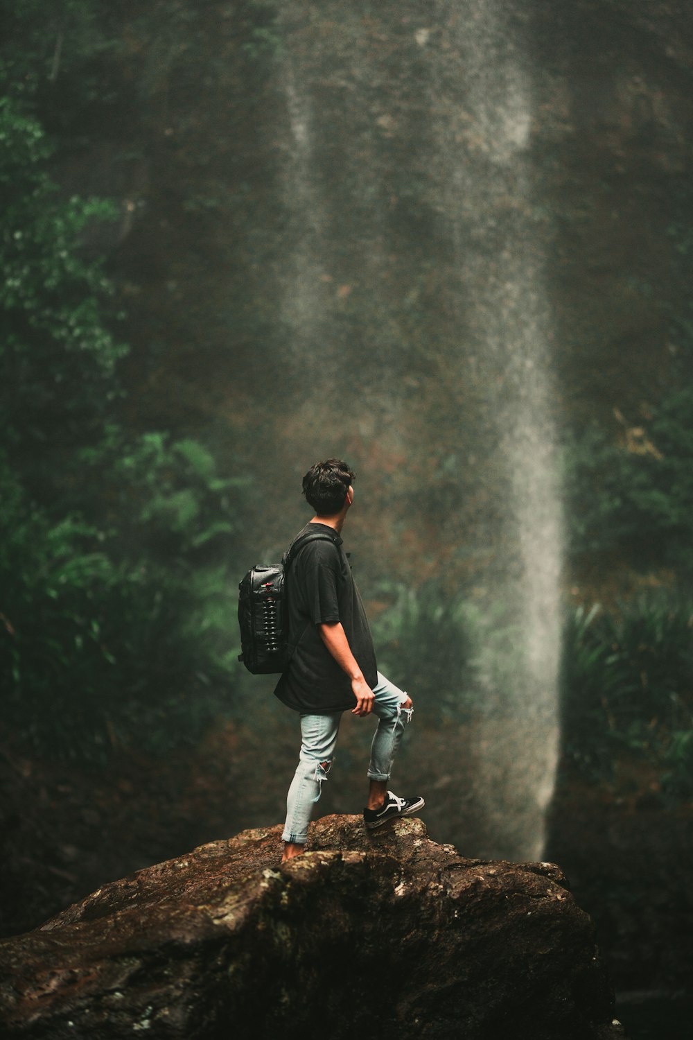 man in black t-shirt and blue denim jeans standing on rock near waterfalls during daytime