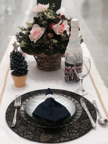 A elegantly set dining table with a decorative plate, navy blue napkin with pearl napkin ring, silver cutlery, and an ornate wine glass. There is a centerpiece featuring pink roses in a basket and a small Christmas tree decoration. A creatively designed wine bottle rests nearby.