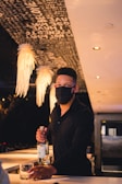 A bartender wearing a black mask stands behind a bar, holding a bottle and a glass. Decorative lighting hangs from the ceiling, and words are visible on the ceiling above.