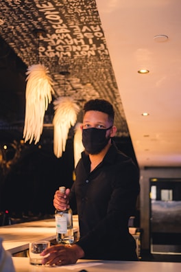 A bartender wearing a black mask stands behind a bar, holding a bottle and a glass. Decorative lighting hangs from the ceiling, and words are visible on the ceiling above.